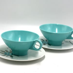 May include: Two turquoise teacups with matching saucers. The saucers have a white background with a black and gray floral design.