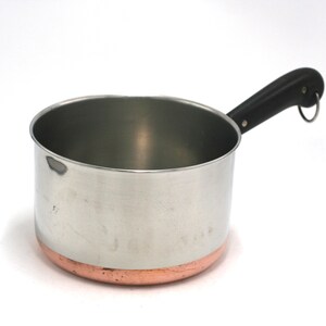 May include: A stainless steel saucepan with a copper-toned base and a black handle. The handle features a small metal loop at the end for hanging. The interior and exterior walls are smooth and metallic, showing a cylindrical shape and a pour spout.