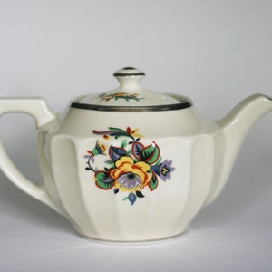 May include: A white teapot with a floral design in shades of yellow, blue, green, and red. The teapot has a silver trim around the lid and a fluted body.