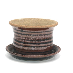May include: A handmade ceramic planter with a matching saucer. The planter is cylindrical with horizontal bands of brown, black, and tan. The saucer is a dark brown color. The planter is suitable for small plants.