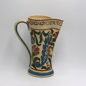 May include: A tall, cream-colored ceramic pitcher with a curved handle and a flared top. The pitcher is decorated with floral and leaf patterns in shades of blue, green, and brown. The base and handle have blue accents.