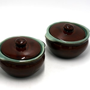 May include: Two ceramic lidded bowls. The bowls are a glossy dark brown with a light green rim. Each bowl has a matching brown lid with a small knob. The bowls are on a white background.