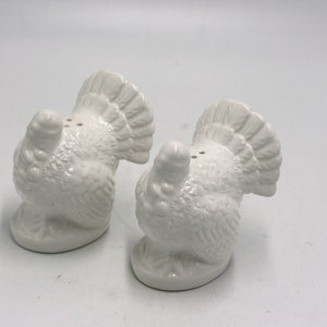 May include: A set of two white ceramic turkey salt and pepper shakers. The turkeys are facing away from each other with their tails up.