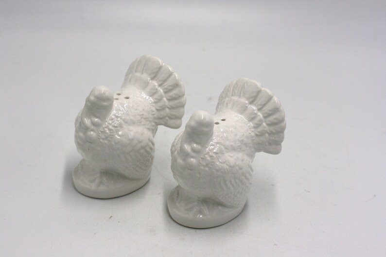 May include: A set of two white ceramic turkey salt and pepper shakers. The turkeys are facing away from each other with their tails up.