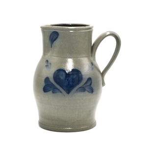 May include: A gray ceramic pitcher with a blue heart and floral design. The pitcher has a handle and a rounded body.