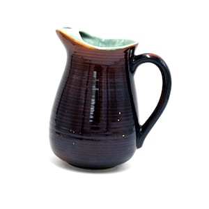 May include: A brown ceramic pitcher with a light green glaze on the inside rim. The pitcher has a rounded body and a handle.
