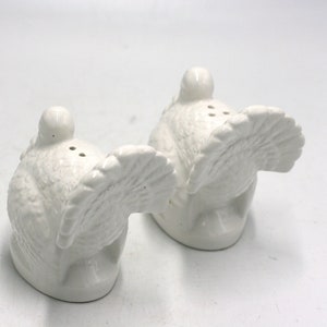 May include: A pair of white ceramic turkey salt and pepper shakers. The turkeys are facing opposite directions with their tails up.