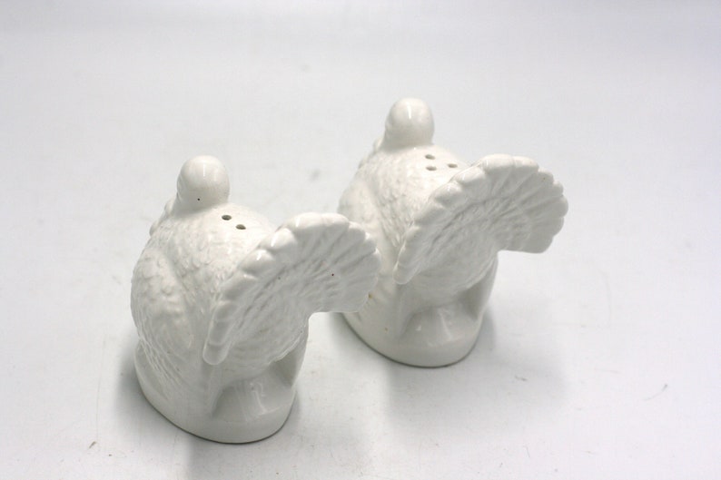 May include: A pair of white ceramic turkey salt and pepper shakers. The turkeys are facing opposite directions with their tails up.