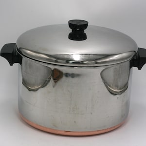 May include: A stainless steel pot with a lid and black handles. The pot has a copper-colored base and a black knob on the lid. This cookware is suitable for cooking.