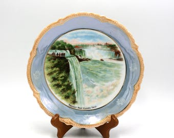 May include: A blue and gold ceramic plate with a painted image of Niagara Falls. The plate has a scalloped edge and a gold rim. The text "Prospect Point Niagara Falls" is printed on the bottom of the plate.