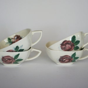 May include: Four white ceramic teacups with a floral design featuring pink roses and green leaves. The cups are stacked in two pairs.