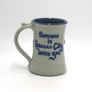 May include: A handmade ceramic mug with a light gray exterior and a dark blue interior. The mug features a curved handle and the blue text "Someone in Kansas City loves you."