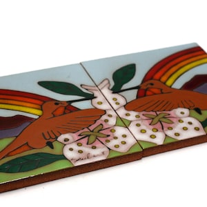 May include: Two ceramic tiles depicting hummingbirds with outstretched wings, perched on a branch with pink flowers. The background features a rainbow and a stylized mountain range. The tiles are signed "Zulbert".