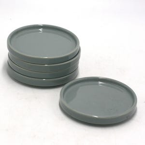 May include: A stack of six gray ceramic coasters, with a seventh coaster placed separately. The coasters are round with a slightly raised edge. The color is a muted, cool gray, and the surface appears smooth and glossy.