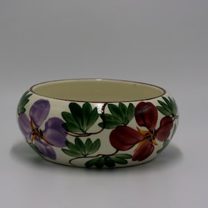 May include: A round, off-white ceramic bowl with a floral design. The bowl features purple and maroon flowers with green leaves. The rim is a dark brown color. The bowl is likely used for serving or display.