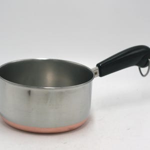 May include: A small stainless steel saucepan with a copper-colored base and a black handle. The handle features a small metal loop at the end for hanging. The interior and exterior walls are metallic silver, while the bottom edge shows a copper band.