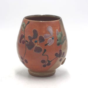 May include: A rounded ceramic cup with a warm orange glaze, featuring hand-painted floral motifs in shades of dark brown, soft pink, and muted green. The vessel has a wide body that tapers toward a circular base and a smooth, open rim.
