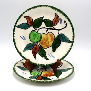 May include: Two white ceramic plates with a green rim and a hand-painted design of apples and leaves. The apples are yellow, red, and green.