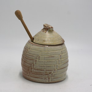 May include: A ceramic honey pot with a lid and a wooden honey dipper. The pot is beige with a brown woven pattern. The lid has a small handle. The honey dipper is inserted into the pot.