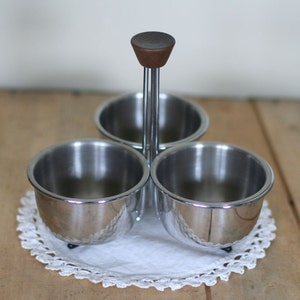 May include: A silver three-section condiment holder with a wooden knob on the top. The holder is sitting on a white crocheted doily.