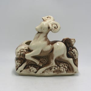 May include: A ceramic figurine of a ram in shades of cream and brown. The ram has large curved horns and is depicted leaping over a textured base. The figurine has a small opening, possibly for a candle or small plant.