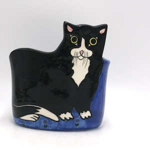 May include: A ceramic cat-shaped container, primarily black with white and pink accents. The cat is depicted with yellow eyes and a white muzzle. The base is blue with floral details. A whimsical and decorative item.