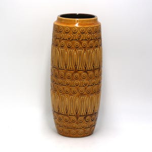 May include: A tall, cylindrical ceramic vase with a brown and tan color scheme. The vase features a repeating pattern of circles and elongated ovals. The interior of the vase is dark brown.