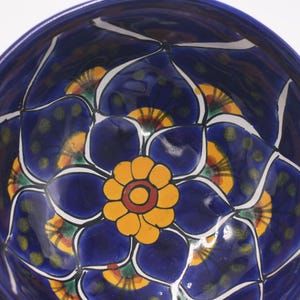 May include: A handcrafted, ceramic bowl with a vibrant floral design. The bowl features a deep blue background with a large yellow flower at the center, surrounded by blue petals and white accents. The bowl's diameter is approximately 10 inches.