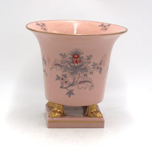 May include: A light pink ceramic vase with a flared rim and gold-toned trim. The exterior features a floral pattern in shades of grey, white, and red. The vase is supported by a square base with four gold-toned feet shaped like animal paws.