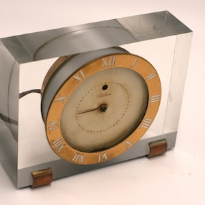 May include: A vintage clock with a white face and black hands, enclosed in a clear acrylic cube with gold accents. The clock is on a white background.