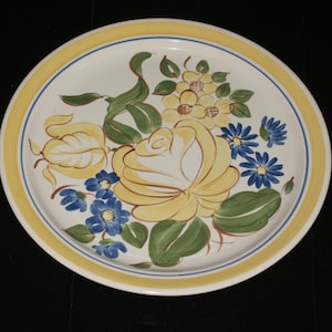 May include: A round, white ceramic plate with a yellow and blue border. The plate features a hand-painted floral design with a large yellow rose, blue flowers, and green leaves. The plate is set against a black background.