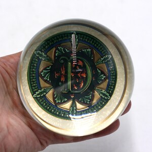 May include: A clear glass paperweight with a detailed design inside. The design features a central letter "D" in green, red, and gold, surrounded by a floral pattern with blue accents. The paperweight is held in a hand.
