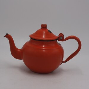 May include: A vintage red teapot with a curved spout, a rounded body, and a matching lid. The handle is arched, and the teapot has a black trim. This classic kitchenware item is likely made of enamel-coated metal.