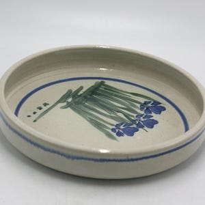 May include: A round, off-white ceramic bowl with a blue rim and a painted floral design. The interior features a cluster of green stems and blue flowers. The bowl is approximately 8 inches in diameter.