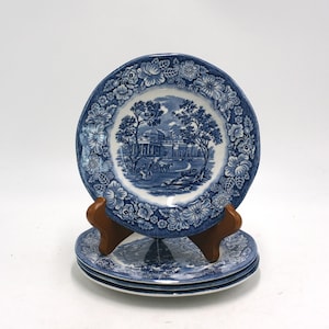 May include: A stack of three round ceramic plates featuring a blue and white transferware pattern. The center depicts a landscape with a building and trees, while the rim is decorated with a detailed floral border. One plate rests on a brown stand.