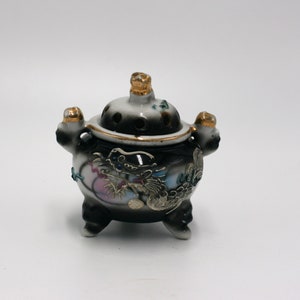 May include: A small, black and white ceramic incense burner with a dragon design and gold accents. The burner has a lid with holes for incense to burn through.