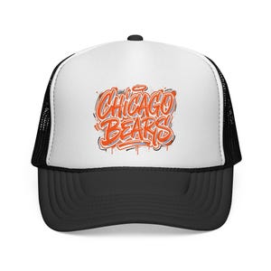 May include: A white and black trucker hat featuring a graphic print on the front panel. The design displays the text CHICAGO BEARS in a stylized orange and white graffiti-inspired font with black outlines and paint splatter accents.