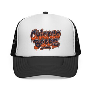 May include: A white and black trucker hat featuring a graffiti-style graphic on the front panel that reads Chicago Bears in orange and navy blue lettering with a dripping paint effect. The cap has a solid black brim and black mesh back panels.