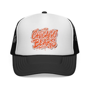 May include: A white and black trucker hat featuring a graphic on the front panel with the text CHICAGO BEARS written in a stylized orange and white graffiti-inspired font with blue outlines and paint splatter effects on a white background.