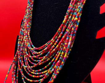 May include: Multi-colored beaded necklace with a layered design. The necklace is made of small, round beads in various shades of red, pink, blue, green, yellow, and white. The necklace is displayed on a black stand against a red background.