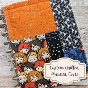 May include: Custom quilted planner cover featuring a patchwork design. The cover includes orange fabric with a constellation pattern, black fabric with white triangles, and a character print. The text "Custom Quilted Planner Cover" is displayed.