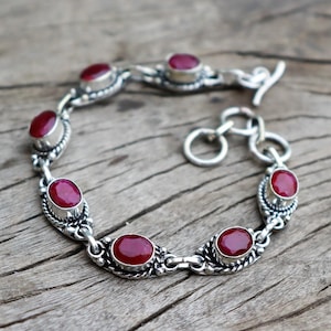 Puede incluir: Un brazalete de plata con cierre de palanca presenta seis piedras preciosas rojas de forma ovalada. El brazalete está sobre una superficie de madera.
