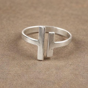 Op de afbeelding: Een zilveren ring met een minimalistisch ontwerp. De ring heeft twee verticale staven die door een band zijn verbonden. De ring is gemaakt van een glanzend, metalen materiaal en staat op een bruine achtergrond.
