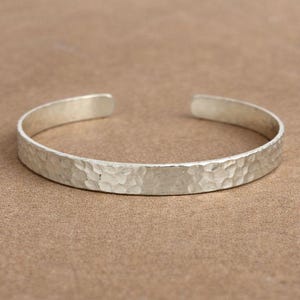 May include: A silver cuff bracelet with a hammered texture. The open-ended bracelet is made of a shiny, light-colored metal. The bracelet is shown on a brown surface.