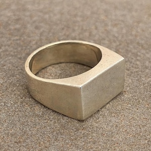May include: A silver-toned ring with a rectangular face. The ring has a thick band and a flat, geometric design. The ring is set against a neutral, textured background.