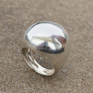 May include: A polished silver ring with a large, rounded top. The ring's surface is smooth and reflective, with a thick band. This statement ring is suitable for everyday wear or special events. The ring is made of silver.