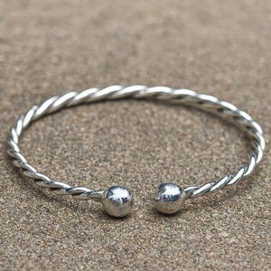 May include: A silver-coloured cuff bracelet with a twisted rope design. The bracelet has two small, round, silver-coloured ball ends. The bracelet is open and sits on a textured, light brown surface.