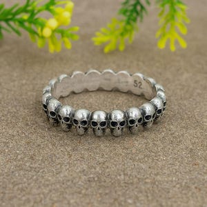 May include: A silver ring with a repeating pattern of small skull designs. The ring has a textured appearance and a size marking of "52" is visible. The ring is displayed on a neutral-toned surface.