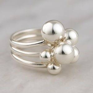 Puede incluir: Un anillo de plata con un grupo de esferas pulidas. El anillo tiene tres bandas y un grupo de esferas de varios tamaños, creando un diseño único y moderno. Las esferas son lisas y reflectantes, añadiendo elegancia.