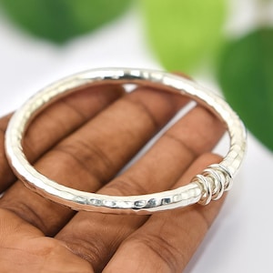Puede incluir: Un brazalete de plata con una textura martillada y una serie de pequeños anillos decorativos. El brazalete es redondo y parece estar hecho de metal. El brazalete se sostiene en una mano.